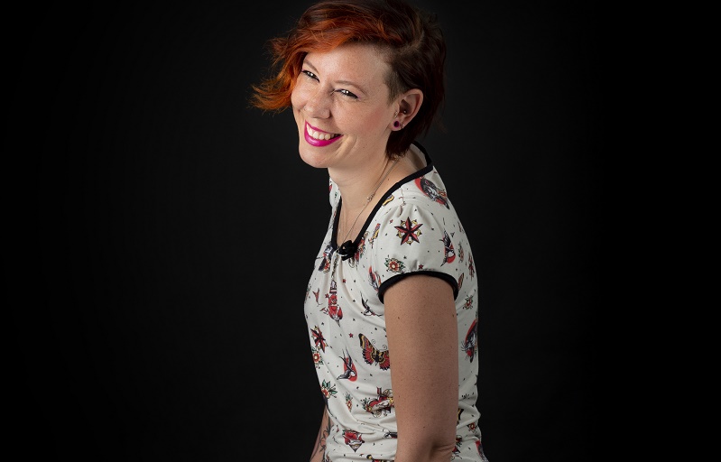 Naomi met kort, goudbruin haar glimlacht naar de camera in een wit shirt met patroon voor een zwarte achtergrond.