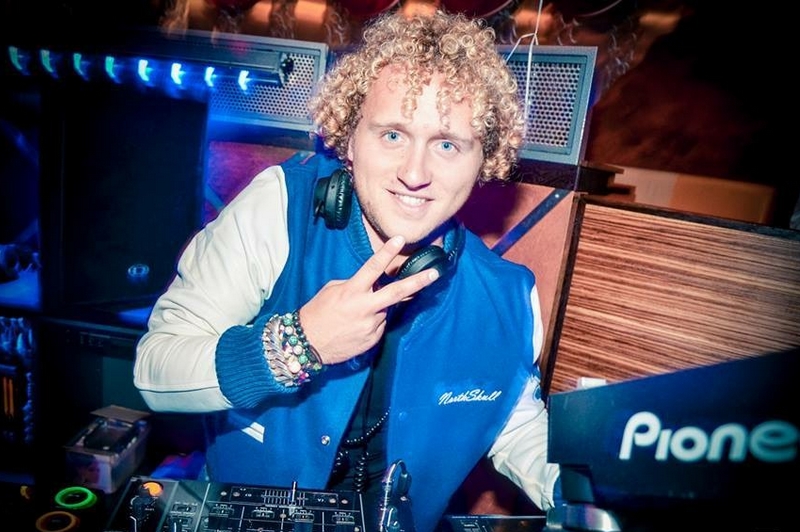 Persoon met krullend blond haar in blauw-wit jasje bij DJ-booth, koptelefoon om nek, geeft peace-teken en lacht naar camera.