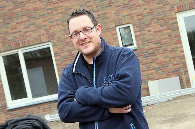 Man met bril en donkerblauwe jas staat met gekruiste armen voor bakstenen gebouw, bouwmaterialen liggen op de grond.