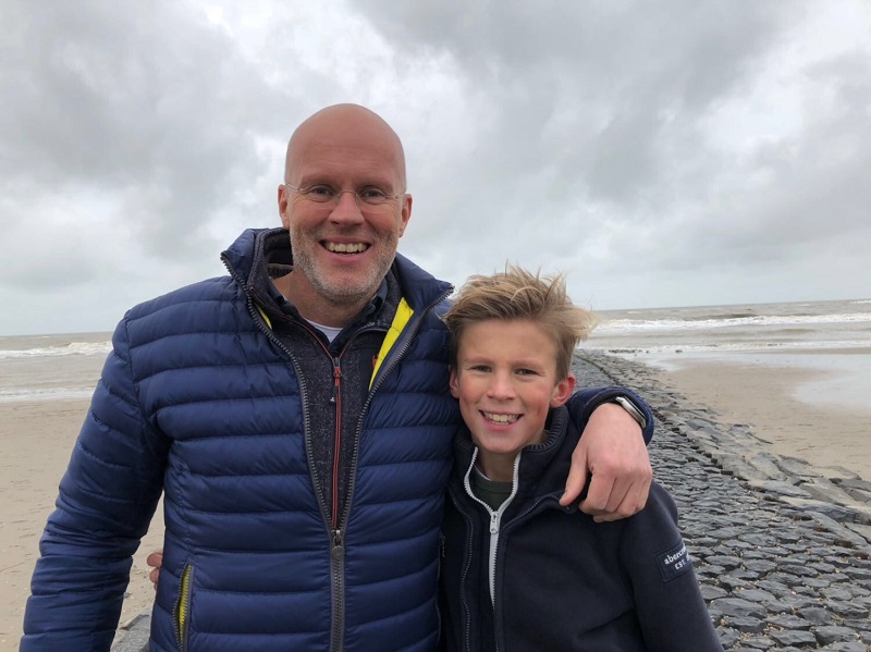 Twee mensen staan samen op een stenen pad aan het strand en lachen naar de camera; volwassene met bril en blauwe jas, vriendelijk erbij.