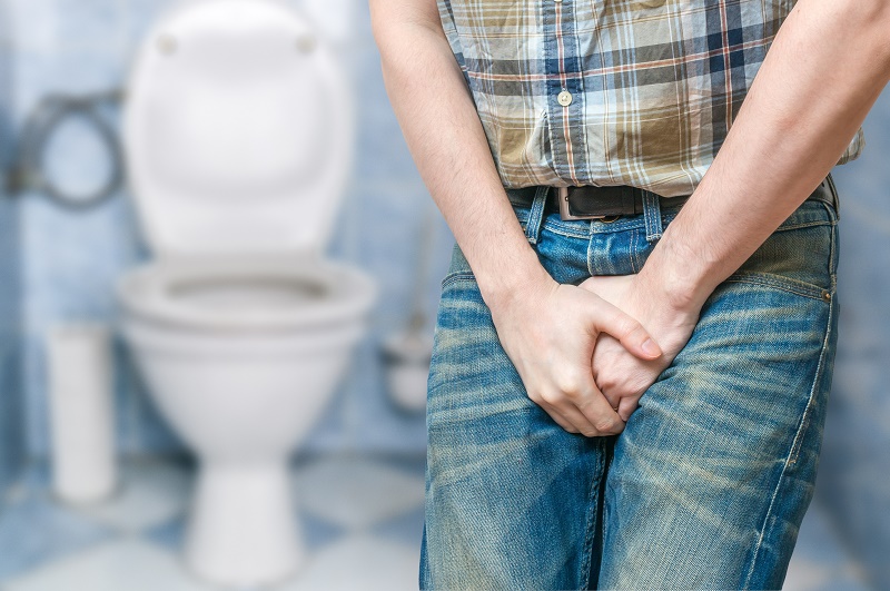 Persoon met geruite blouse en spijkerbroek houdt handen bij kruis voor toilet; zichtbaar ongemak in badkamer.