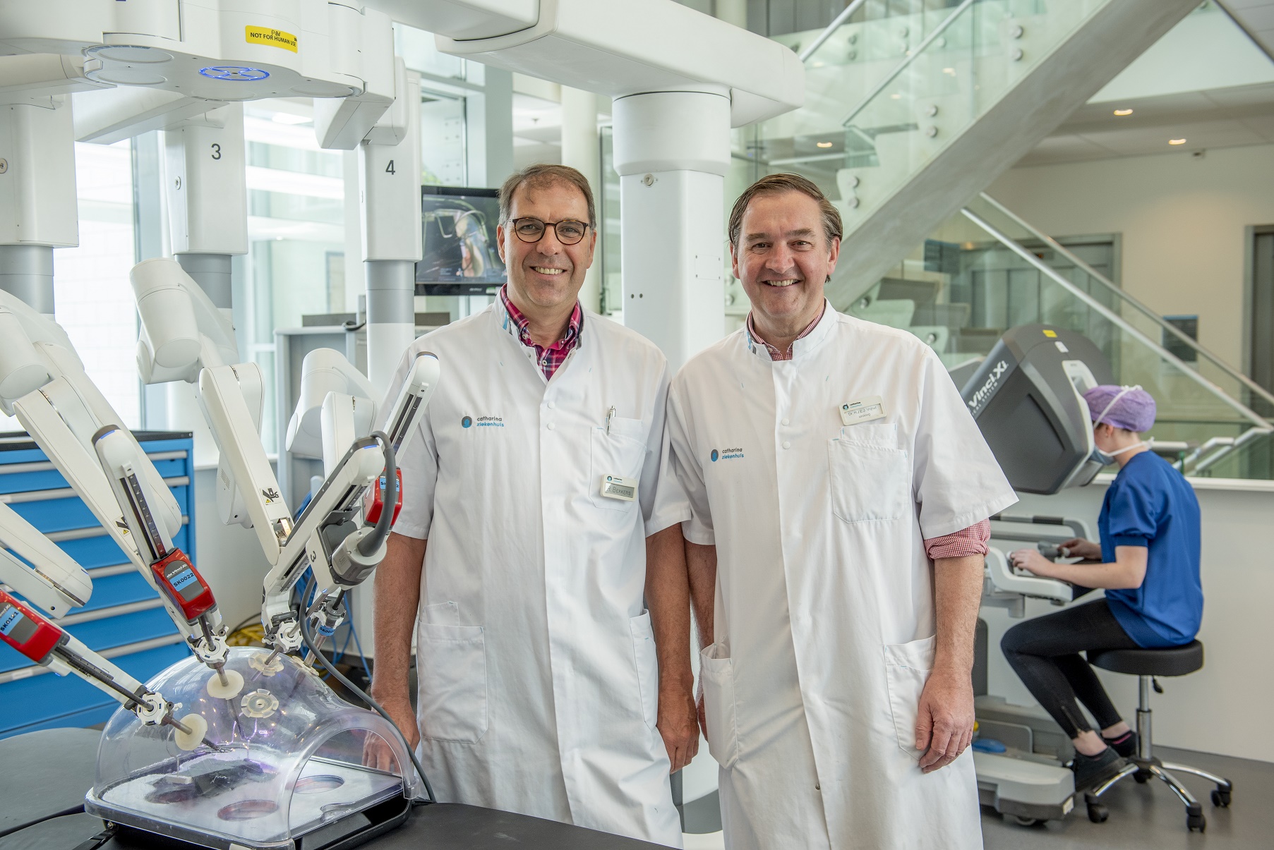 Twee mannelijke dokters in witte jassen bij robotchirurgie-apparatuur; persoon in blauwe scrubs aan console, helder verlicht lab.