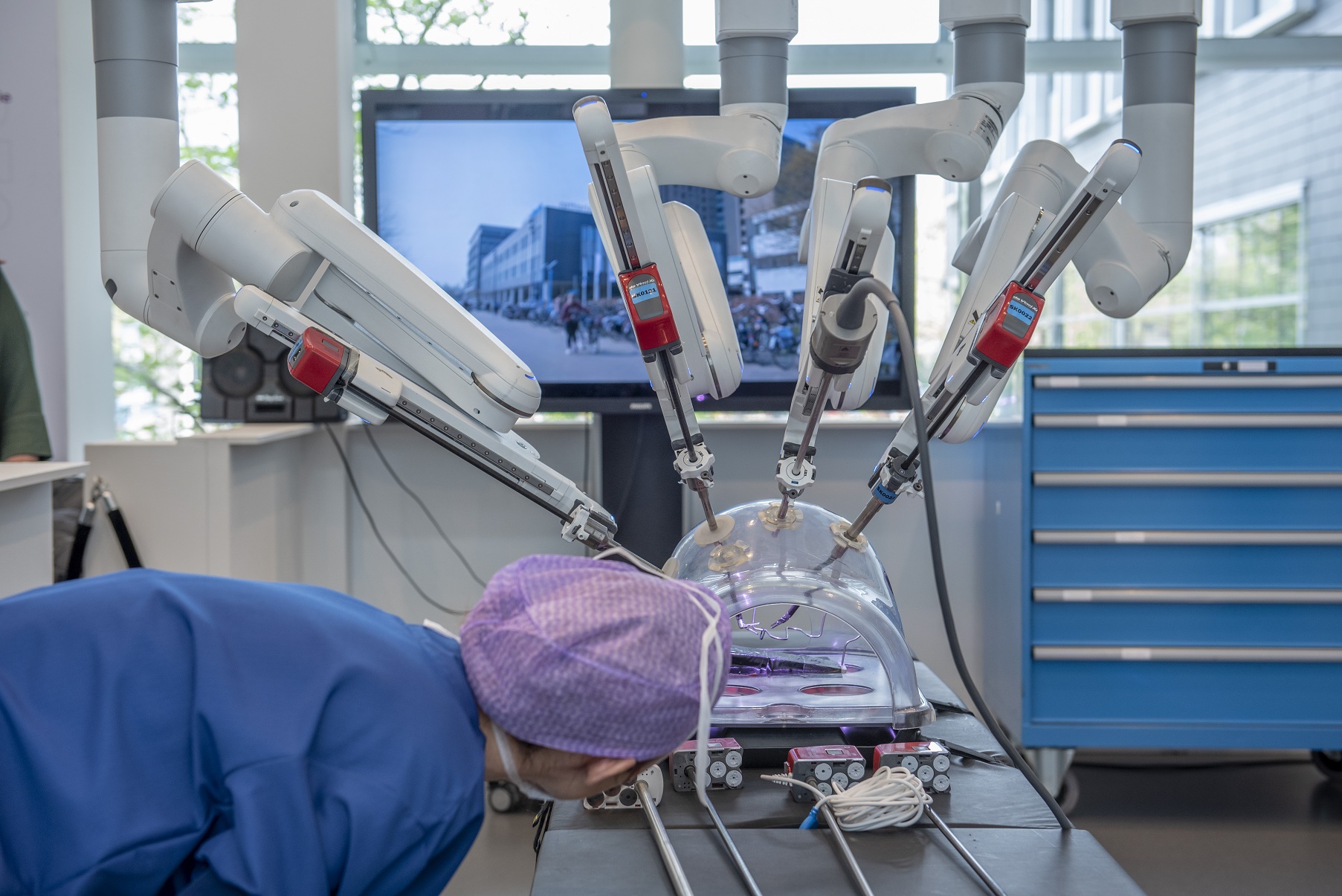Persoon in OK-kleding bekijkt operatierobot met meerdere armen, die op een model werkt; procedure is te zien op monitor.