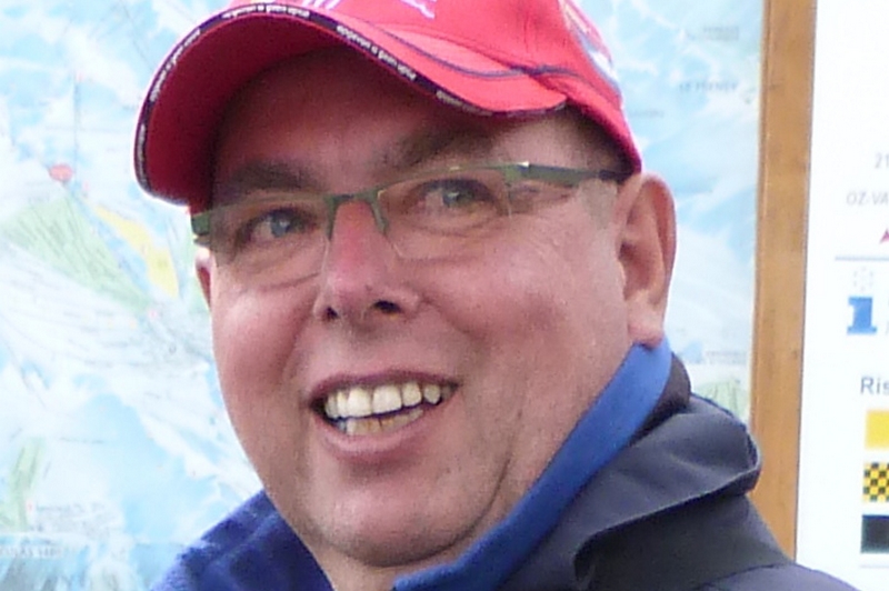 Een man met bril, rode pet en blauwe jas lacht buiten; op de achtergrond is een vage kaart te zien.
