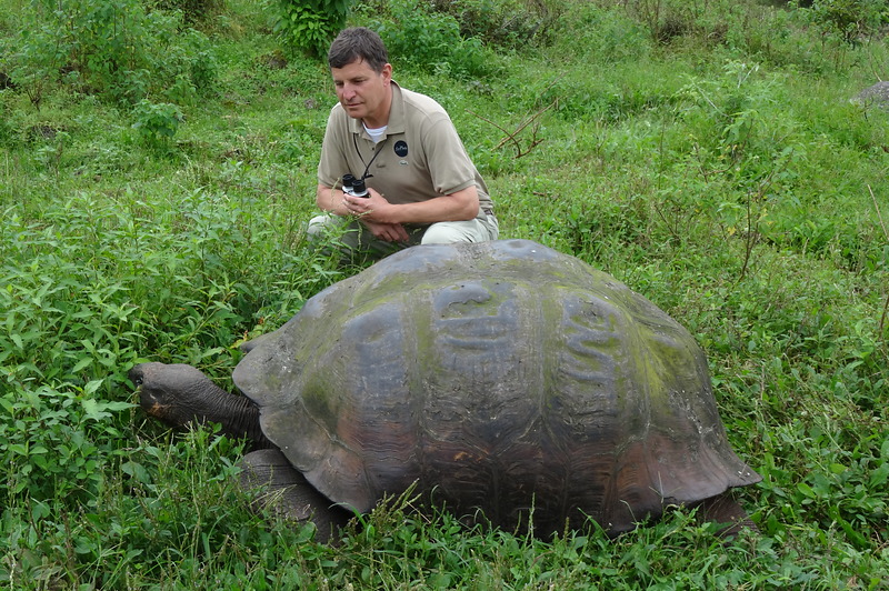 Een cardioloog in beige kleding hurkt in het gras naast een groot schildpad in een groene, weelderige buitenomgeving.