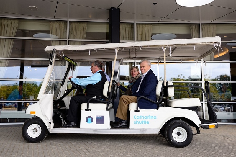 Een man bestuurt een witte Tweede Catharina shuttle met twee oudere passagiers, geparkeerd bij een gebouw met grote ramen.