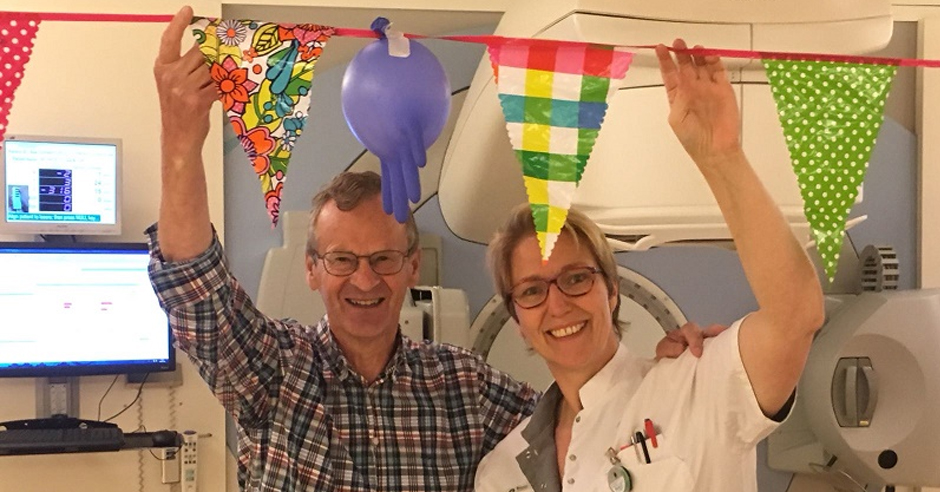 Twee mensen staan binnen onder gekleurde vlaggetjes, één in ruitjesblouse, één in witte jas; medische apparatuur zichtbaar.
