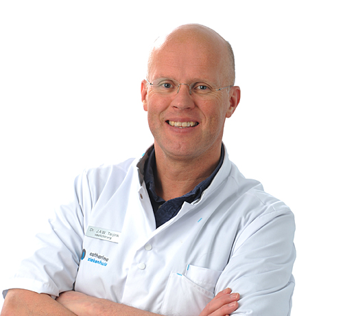 Persoon met bril en kaal hoofd draagt een witte labjas en lacht met gekruiste armen. De achtergrond is effen wit.