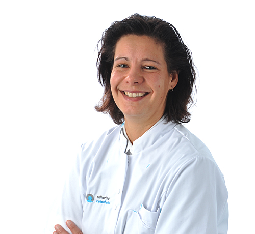 Persoon met kort donker haar lacht en staat met gekruiste armen in een witte doktersjas tegen een witte achtergrond.