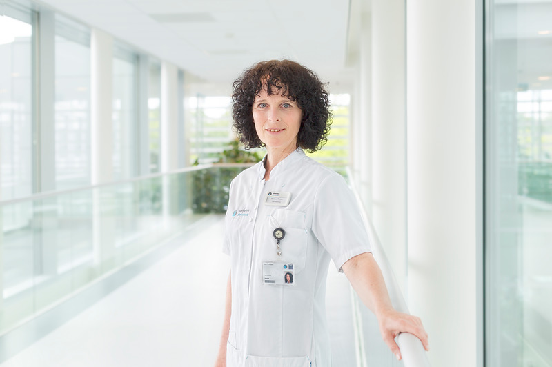 Vrouw in witte medische jas met badge staat in lichte bende, hand op leuning. Ze kijkt in de camera. Zonlicht door ramen.