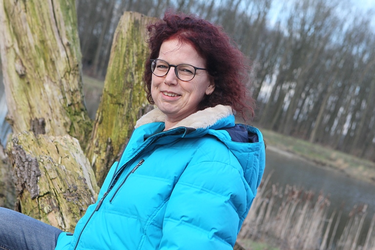 Vrouw met bril en felblauwe winterjas zit op boomstam bij vijver, omringd door boerenkool bomen.