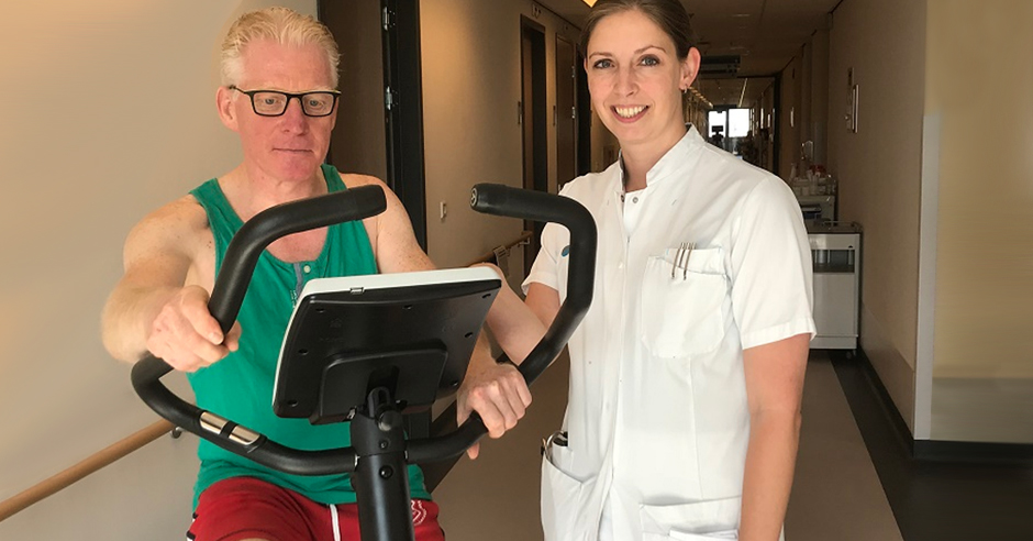 Man op hometrainer in gang, draagt groene tanktop en rode short; verpleegkundige helpt, lacht naar camera.