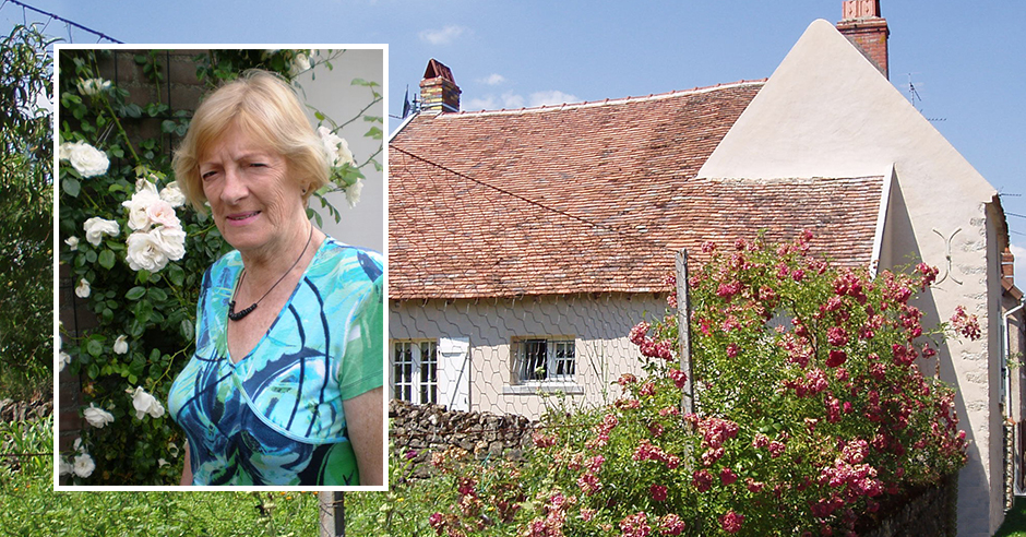 Vrouw met kort blond haar bij witte rozen in Franse tuin, blauwe top, oud huis met pannendak, roze bloemen, blauwe lucht.