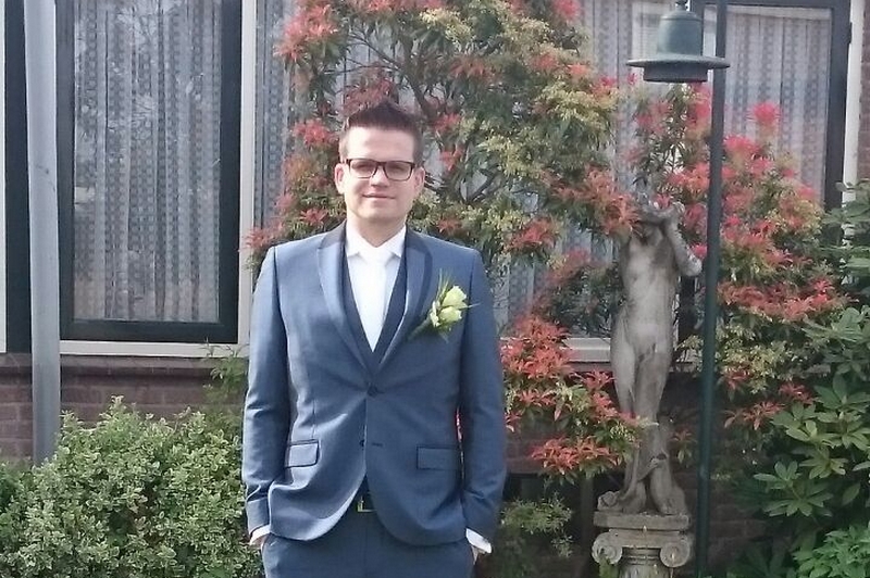 Persoon in blauw pak met witte blouse en corsage poseert buiten bij struik, beeld en gebouw met grote ramen.