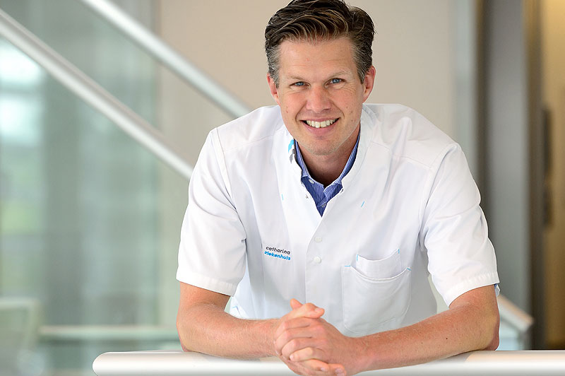 Man met witte doktersjas leunt glimlachend op een reling. Binnen, met vage glas- en metaalelementen op de achtergrond.