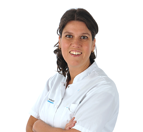 Vrouw met donker haar in een wit medisch uniform staat met gekruiste armen en glimlacht voor een witte achtergrond.