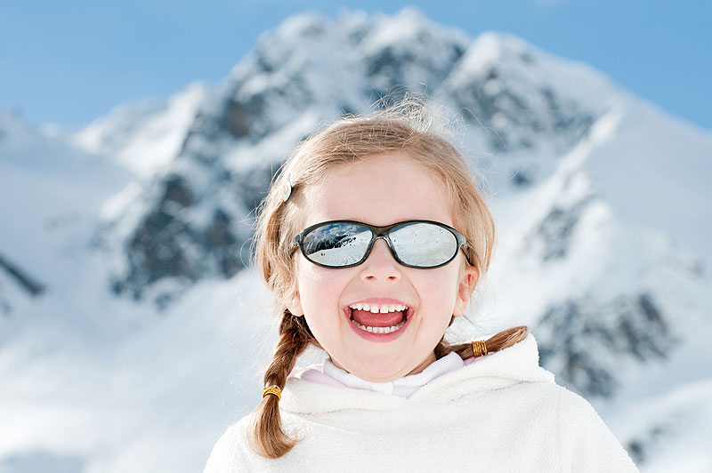 Kind met vlechtjes, zonnebril en witte jas lacht voor besneeuwde bergen onder een blauwe lucht, beschermd tegen felle zon.