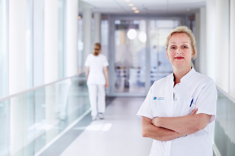 Een borstkankerchirurg in wit uniform staat met gekruiste armen in een lichte bende. Op de achtergrond loopt iemand weg.