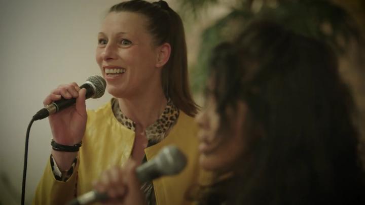 Twee vrouwen zingen met microfoons; één in gele jas staat scherp op de foto, de ander vaag. Er is een plant zichtbaar.