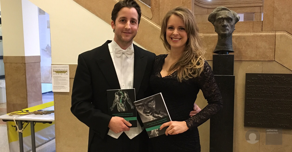 Man in smoking en vrouw in zwarte jurk staan binnen met elk een boek. Achter hen een bronzen buste op sokkel, promotiebanners.