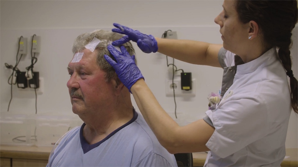 Een zorgverlener met paarse handschoenen onderzoekt de hoofdhuid van een oudere man met verband om het hoofd, in een kliniek.