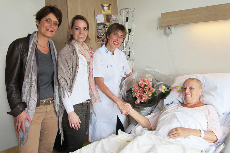 Patiënt in ziekenhuisbed krijgt bloemen en handdruk van verpleegkundige op Dag van de Eierstokkanker; anderen glimlachen.