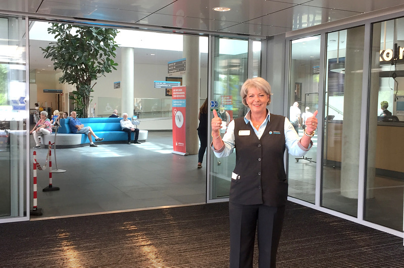 Vrouw in uniformvest geeft twee duimen omhoog bij schuifdeuren. Op de achtergrond mensen op blauwe banken in moderne lobby.