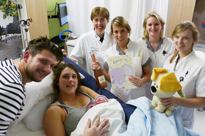 Vrouw in kraamsuite op ziekenhuisbed houdt haar eerste baby vast, omringd door man en vier lachende verpleegkundigen.