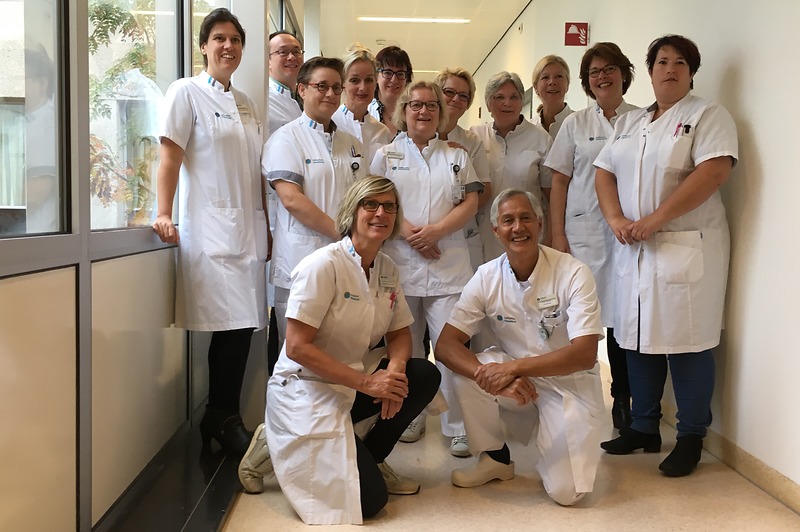 Dertien zorgprofessionals in witte jassen, sommigen met keurmerkbadge, poseren in een lichte bende. Twee knielen vooraan.