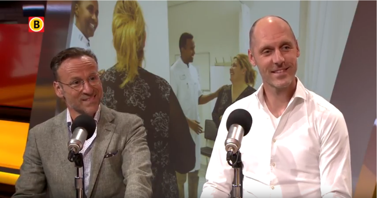Twee mannen zitten met microfoons aan tafel voor een uitzending. Op het scherm achter hen zie je mensen lachen tijdens een etentje.