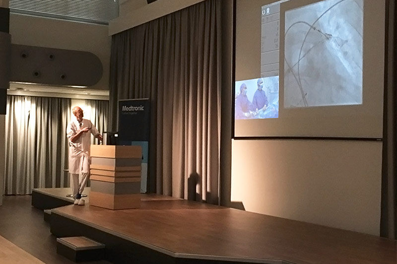 Een persoon in witte jas geeft een presentatie in het Catharina Ziekenhuis, met medische beelden en live Hartweek-chirurgie op scherm.