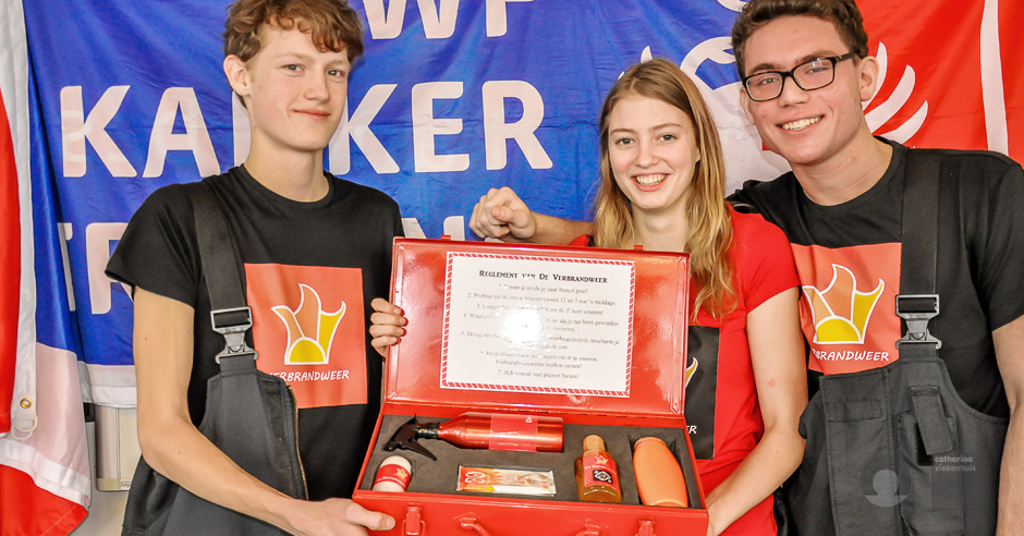 Drie jongeren met shirts met kroonlogo tonen lachend een rode koffer met brandblusser en certificaat bij Catharina Ziekenhuis.