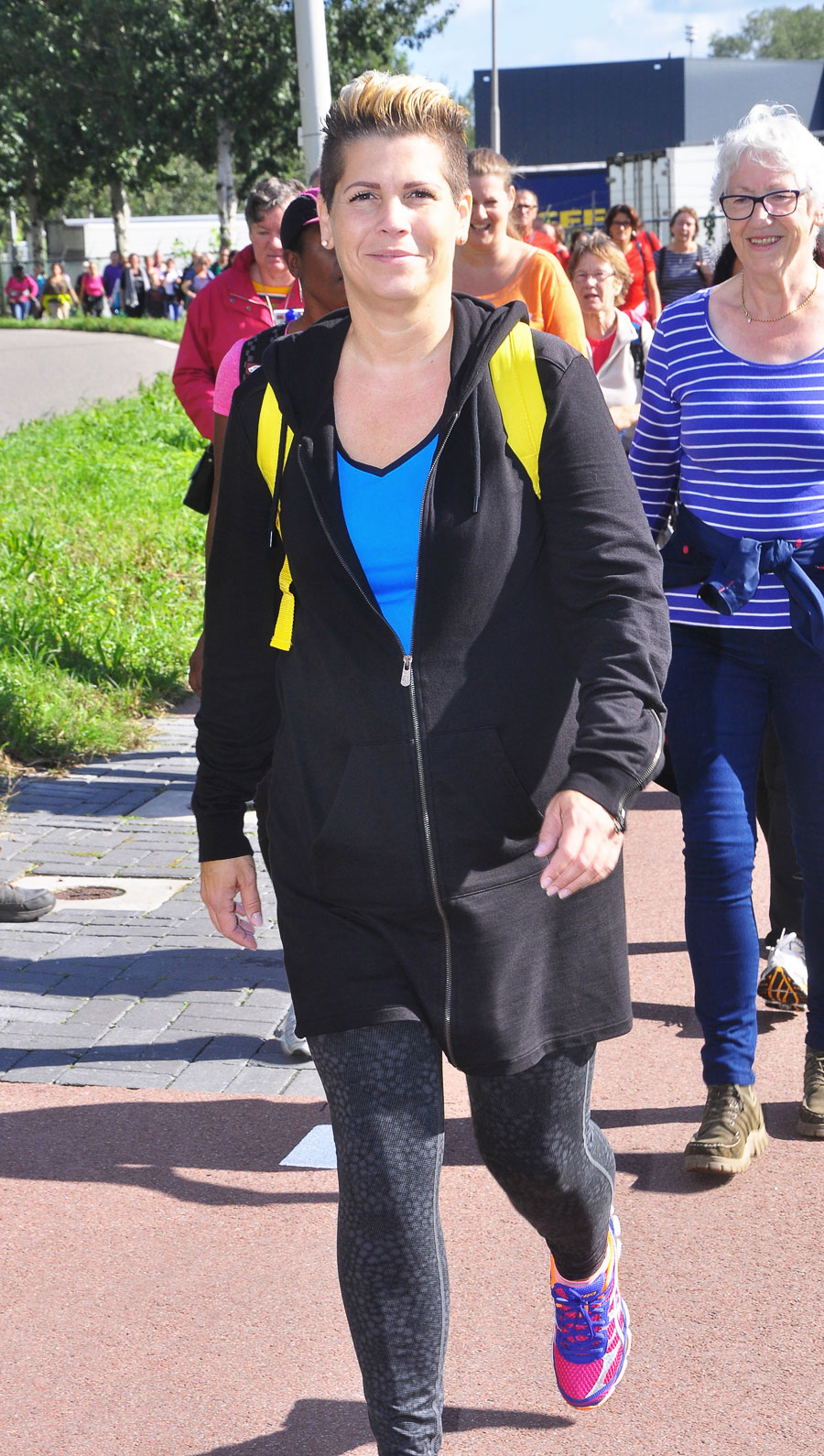 Een patiënt met kort haar, zwarte jas, blauw shirt en roze schoenen loopt buiten. Achter hen loopt een groep mensen.