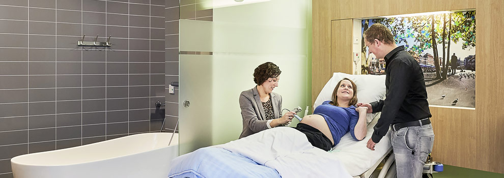Zwangere vrouw op nieuw kraambed krijgt echo; partner houdt haar hand vast. Bad en tegelmuur tonen moderne faciliteiten.