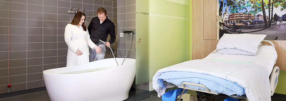 Zwanger vrouw en man bij bevalbad in betegelde kraamsuite. Naast hen een opgemaakt ziekenhuisbed en een schilderij aan de muur.
