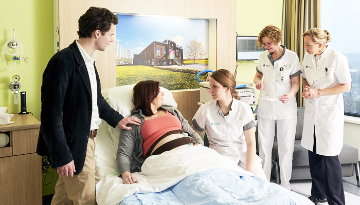 Zwangere vrouw in ziekenhuisbed met monitoring, man erbij, drie vrouwelijke zorgverleners in witte kleding in nieuwe kraamsuites.