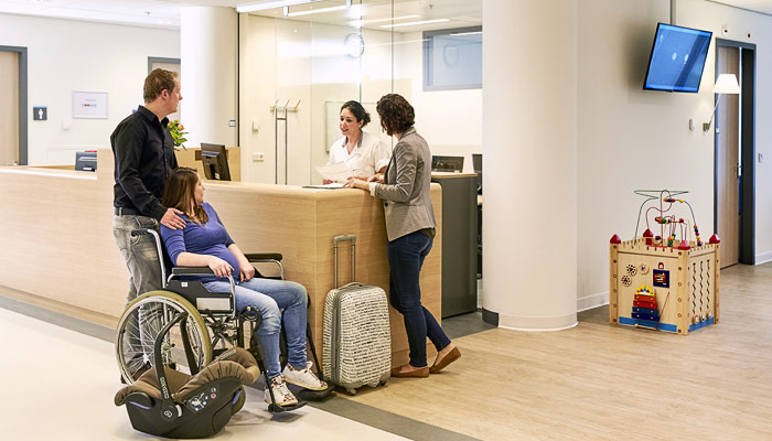 Drie volwassenen bij een ziekenhuisbalie, vrouw in rolstoel met koffer. Op de achtergrond kraamsuites en speelhoek.