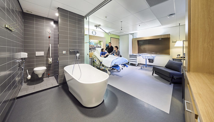 Een nieuwe kraamsuite met bad, privébadkamer links, centraal bed met patiënt en drie verzorgers; moderne apparatuur actief.