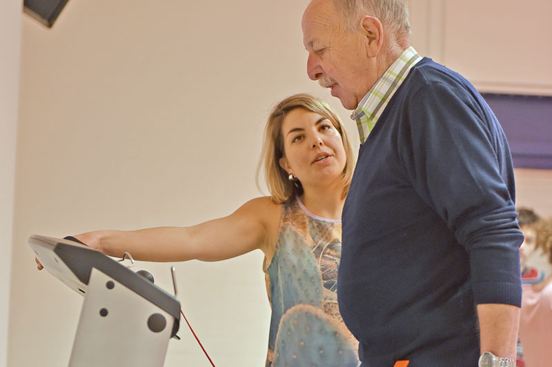 Vrouw wijst naar loopbandpaneel en spreekt met oudere man tijdens gesuperviseerde looptraining in neutrale binnenruimte.