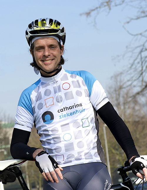 Wielrenner Lukas Dekker in blauw-wit Catharina Ziekenhuis-shirt buiten, met één hand op zijn fiets. Bomen en heldere lucht achter hem.