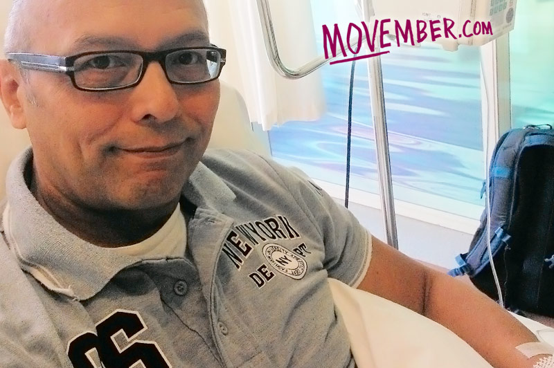 Man met bril en grijs poloshirt zit met infuus in arm in medische ruimte. MOVEMBER.com staat in roze op de achtergrond.