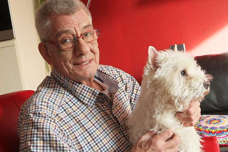 Oudere man met bril en geruite blouse zit binnen, glimlacht met een wit hondje op schoot. Rode muur en bank op achtergrond.