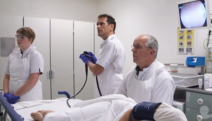 Drie artsen voeren in een ziekenhuis een endoscopie uit voor darmkankeronderzoek; medische apparatuur op de achtergrond.