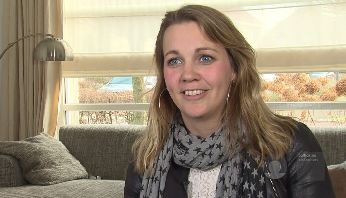 Blonde vrouw met sjaal met sterren zit glimlachend op grijze bank in lichte woonkamer in Eindhoven met neutraal interieur.