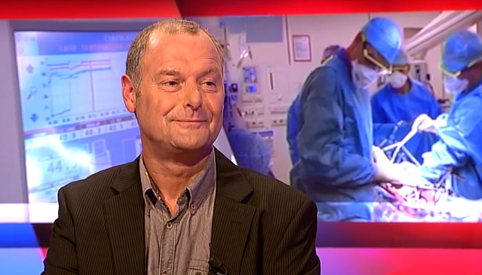 Man in pak zit in studio; op groot scherm achter hem artsen die opereren bij uitgezaaide darmkanker, onderwerp overlevingskans.