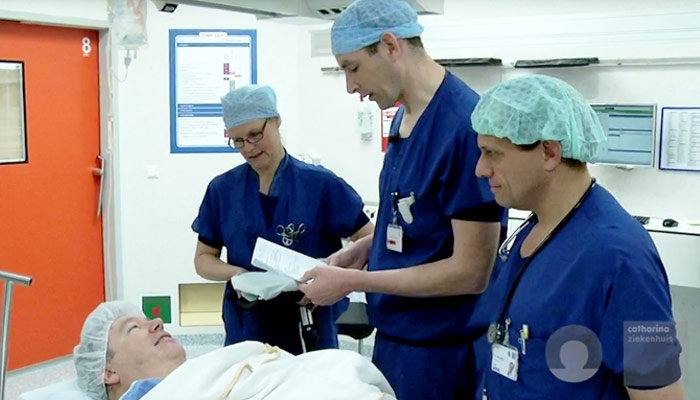 Drie artsen in blauwe scrubs staan bij een patiënt in bed en bespreken documenten over een gastric sleeve in een lichte kamer.