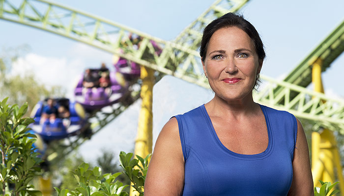 Vrouw in blauwe top buiten, lachend naar de camera. Op de achtergrond een achtbaan met groene rails en gekleurde karretjes.