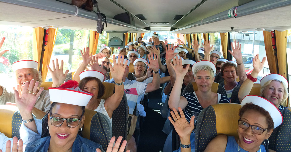 Vrijwilligers met witte hoeden en rode rand zitten glimlachend in een bus met gele en grijze stoelen, zonlicht schijnt binnen.