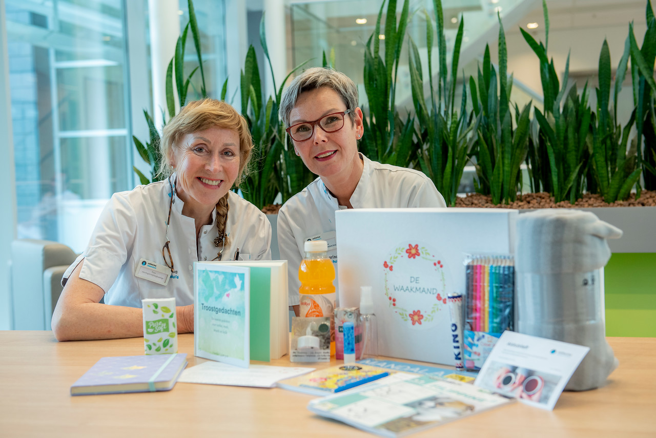 Twee vrouwen in witte uniformen zitten aan tafel met een mand vol troost. Op de achtergrond staan hoge groene planten.