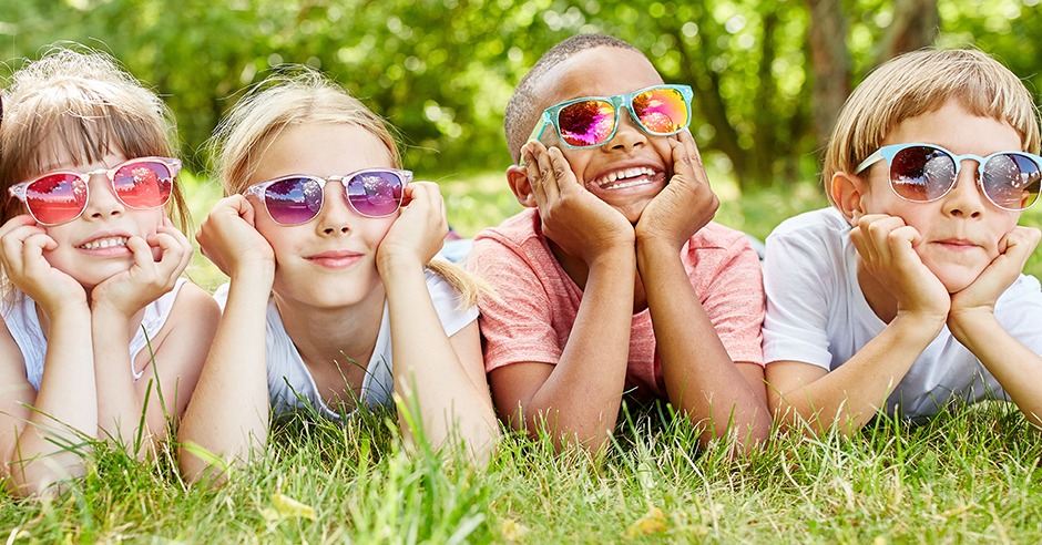 Vier kinderen met zonnebrillen liggen op hun buik in het gras. Op de achtergrond zijn bomen en groen zichtbaar.